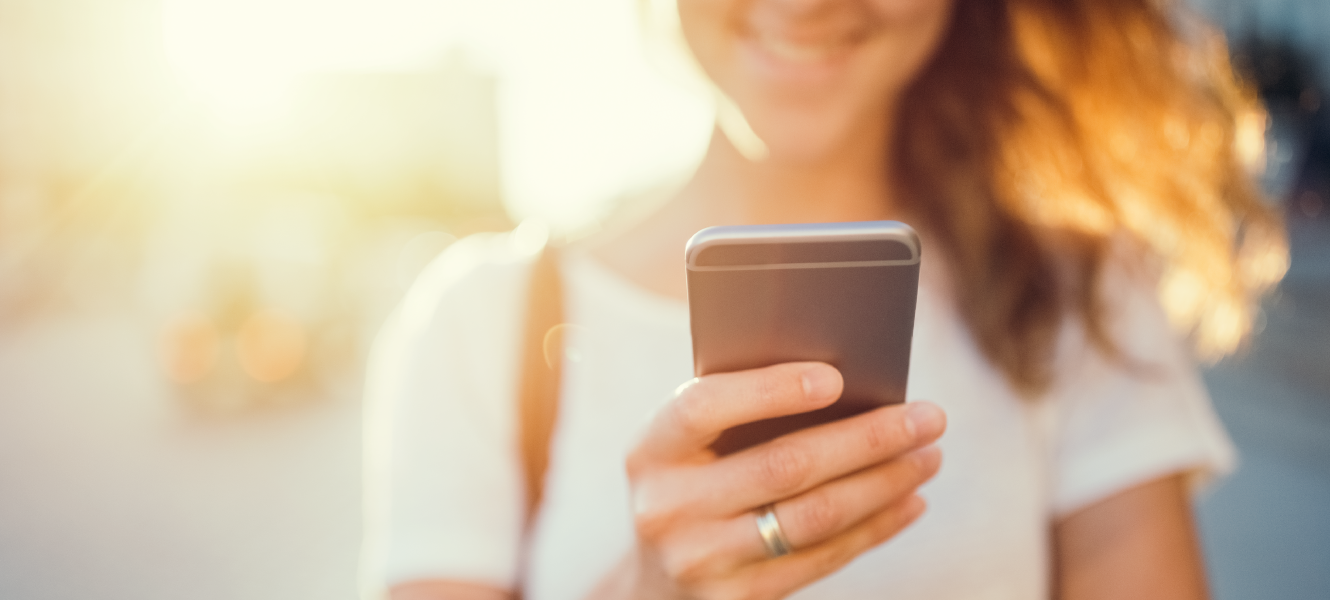 a woman with long brunette hair and wearing a short-sleeve t-shirt smiles while looking at her iphone with the sun behind her