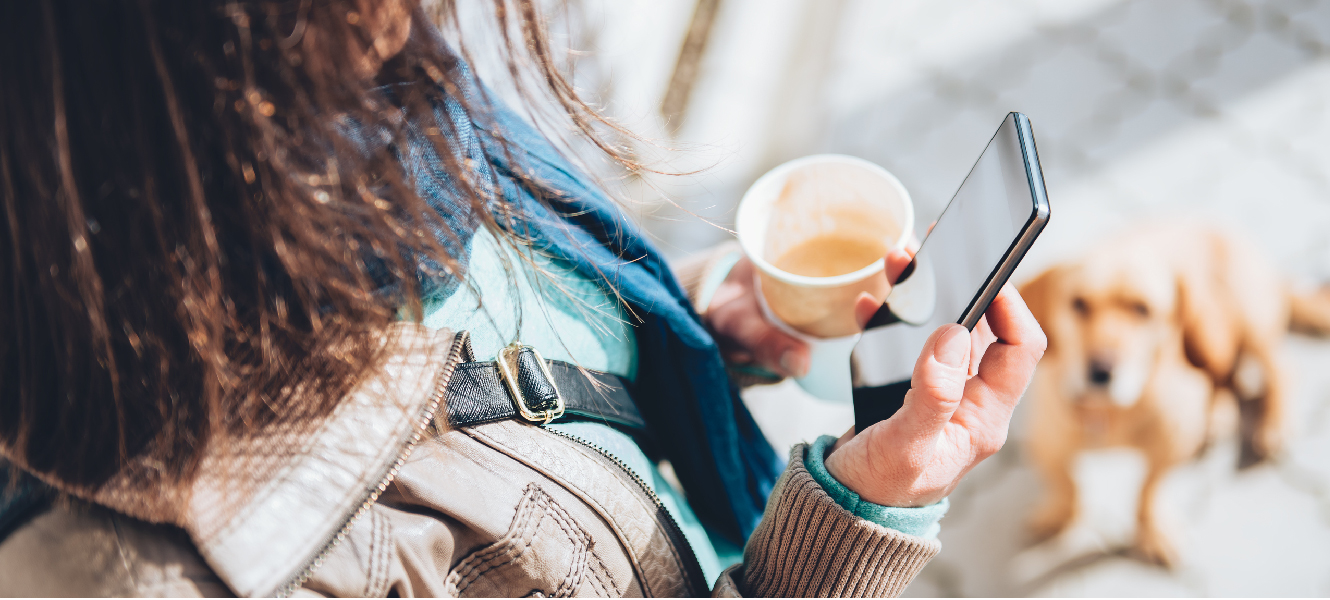 woman holding open coffee cup while looking at smart phone with small dog